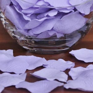 May include: A glass bowl filled with lavender rose petals. The petals are scattered on a wooden surface.