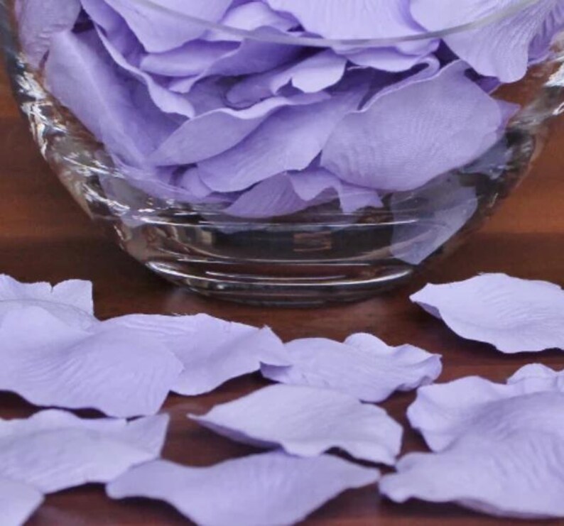 May include: A glass bowl filled with lavender rose petals. The petals are scattered on a wooden surface.