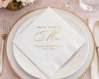 May include: A square white paper napkin featuring gold foil text that reads FROM MISS TO Mrs. Carmen's Bridal Shower 09.17.2022. The napkin has a textured border and is placed on a white plate with a gold beaded rim, accompanied by a silver fork.