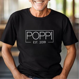 May include: Black t-shirt with the word "POPPI" in a white rectangular frame, with "EST. 2019" below. The shirt is a crew neck style.