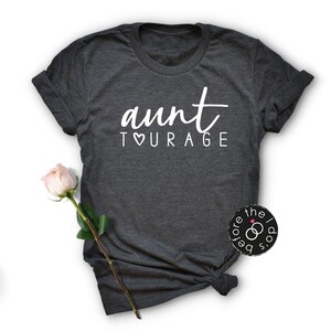 May include: A dark charcoal gray short-sleeve t-shirt featuring the white text "aunt TOURAGE" with a heart symbol replacing the letter O. The shirt has a crew neckline and rolled sleeves, with a single pale pink rose placed to the left side.