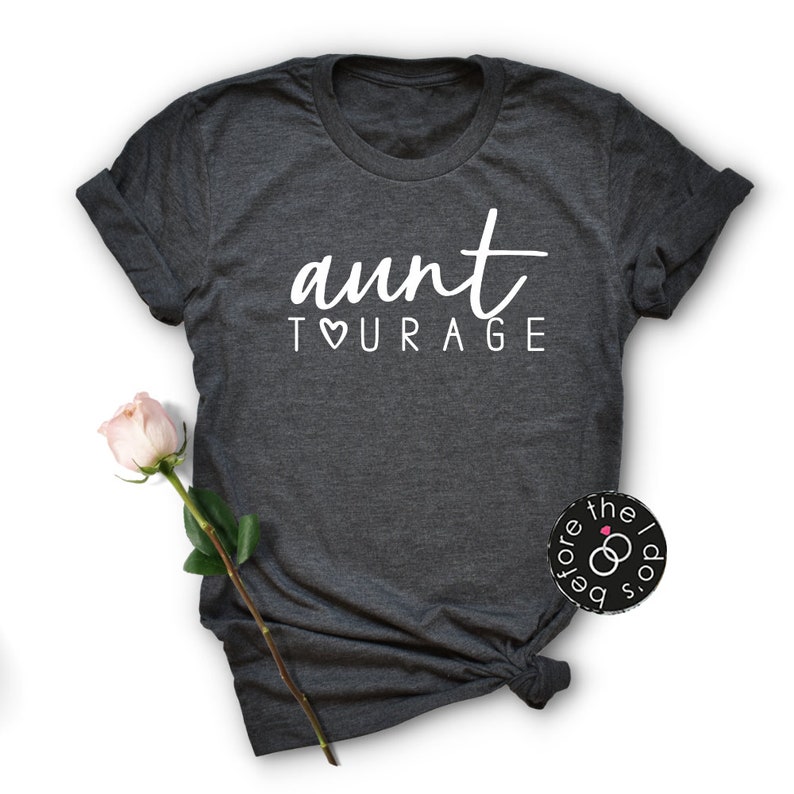 May include: A dark charcoal gray short-sleeve t-shirt featuring the white text "aunt TOURAGE" with a heart symbol replacing the letter O. The shirt has a crew neckline and rolled sleeves, with a single pale pink rose placed to the left side.