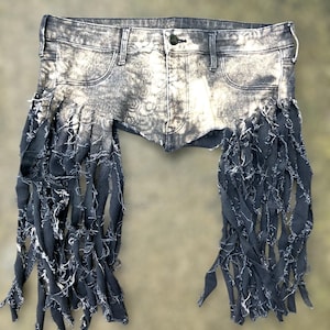 May include: Acid wash denim shorts with distressed detailing and long, fringed accents. The shorts feature a button closure and classic five-pocket design. The fringe is a dark, contrasting color.