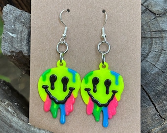 May include: A pair of neon green smiley face earrings with black eyes and mouths. The bottom of the faces have colorful drips in pink, blue, and green. The earrings hang from silver-colored fish hook ear wires.