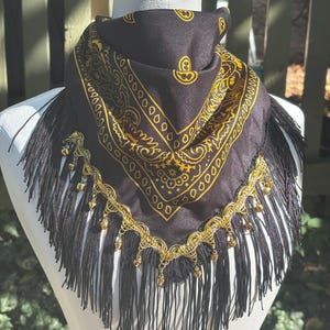 May include: A black bandana featuring a gold paisley pattern, adorned with a decorative gold-toned trim and small metallic bells along the edge, finished with long black fringe hanging from the bottom of the fabric triangle.