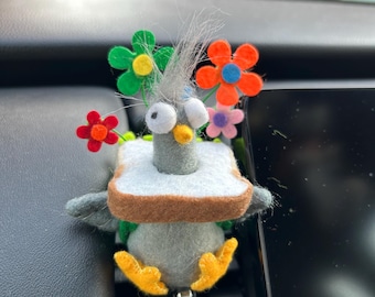 May include: A needle-felted gray pigeon figurine wearing a slice of white bread with a brown crust around its neck. The pigeon has large white eyes, a yellow beak, and is decorated with small felt flowers in green, orange, and purple colors.