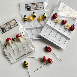 May include: A set of colourful, handmade needlepoint pins featuring ladybirds, bees, and mushrooms. The pins are packaged on white cards with the text "The Snouty Dragon" and a website address.