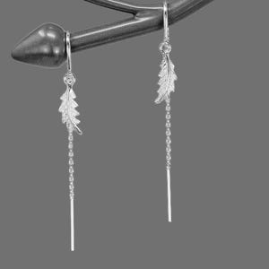 Puede incluir: Un par de pendientes colgantes plateados. Cada pendiente presenta un dije en forma de hoja y una delicada cadena que termina en una barra recta. Los pendientes se muestran sobre un fondo gris.