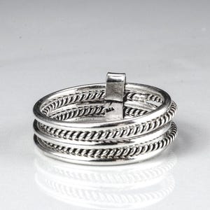 May include: A silver ring with a layered design. The ring features multiple bands, some with a twisted rope texture, and a vertical rectangular accent. The ring is displayed on a white surface.