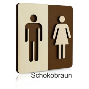 Puede incluir: Un letrero de madera con un s&iacute;mbolo masculino y un s&iacute;mbolo femenino, separados por una l&iacute;nea vertical. El s&iacute;mbolo masculino est&aacute; a la izquierda y el s&iacute;mbolo femenino a la derecha. El letrero est&aacute; pintado en dos colores: madera clara a la izquierda y marr&oacute;n oscuro a la derecha. El texto "Schokobraun" est&aacute; impreso debajo del letrero.