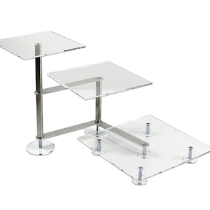 Może przedstawiać: Trójpoziomowy stojak na ciasto z akrylu z srebrną metalową ramą. Stojak ma trzy kwadratowe poziomy i jest podtrzymywany przez cztery srebrne metalowe nogi.