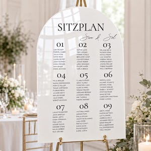 Könnte beinhalten: Ein bogenförmiges Sitzplanschild aus weißem Acryl für Hochzeiten mit dem schwarzen Text SITZPLAN und Sara & Sol, unterteilt in neun nummerierte Spalten mit in schwarzer Tinte gedruckten Namen, auf einem goldenen Metallständer.