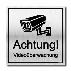 Könnte beinhalten: Ein silbernes Metallschild mit schwarzem Rand zeigt ein schwarzes Überwachungskamera-Symbol. Darunter sind die Worte "Achtung!" und "Videoüberwachung" in Schwarz gedruckt. Das Schild hat eine gebürstete Metallstruktur.
