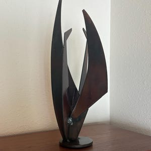 May include: A vertical metal sculpture featuring three distinct, elongated, curved blades of dark, weathered steel. The pieces are welded together at a central point and rise from a circular base, creating an abstract, upward-reaching form.