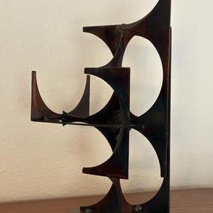May include: A dark brown metal sculpture with an abstract design. The sculpture features curved and geometric shapes, including arches and rectangular elements. It stands on a circular base and is displayed on a wooden surface.