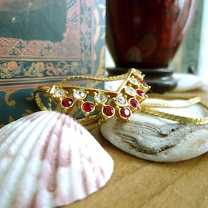 May include: A gold chain necklace with a red and clear crystal pendant in the shape of a V. The necklace is lying on a seashell and a piece of driftwood.