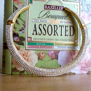 May include: A gold-toned, rhinestone-studded collar necklace. The necklace is in front of a green and floral tea box with the words "BASILUR Bouquet ASSORTED" printed on it. The tea box also lists "Exclusive Green Tea Collection".