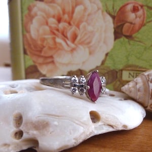 May include: A silver ring with a marquise-cut, deep red gemstone as the focal point. The ring is flanked by smaller, clear gemstones. The ring is displayed on a white, textured surface, with a floral background.