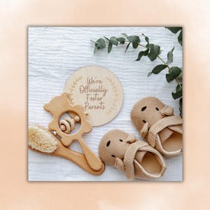 May include: A wooden baby rattle, a soft brush, and a pair of beige baby shoes are arranged on a white surface. A round wooden sign with the text "We're Officially Foster Parents" is also present. Eucalyptus leaves add a natural touch.
