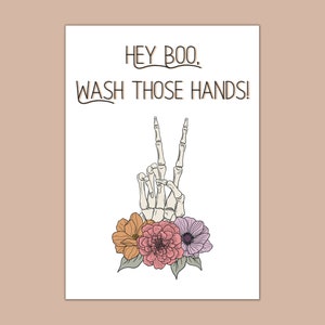 Puede incluir: Una mano de esqueleto blanca con dos dedos levantados en se&ntilde;al de paz, rodeada de flores rosas, naranjas y moradas. El texto "Hey Boo, Wash Those Hands!" est&aacute; escrito encima de la mano.