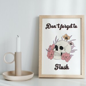 Puede incluir: Una impresi&oacute;n enmarcada con fondo blanco y una calavera en blanco y negro con flores rosas y amarillas. El texto "No olvides tirar de la cadena" est&aacute; escrito en letras cursivas negras.