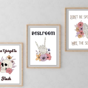 Puede incluir: Tres impresiones enmarcadas con ilustraciones florales y de esqueletos y texto. La primera impresi&oacute;n dice "No olvides tirar de la cadena" con una calavera y flores. La segunda impresi&oacute;n dice "Aseo" con una mano de esqueleto y flores. La tercera impresi&oacute;n dice "No seas espeluznante, &iexcl;limpia el asiento!" con una caja tor&aacute;cica de esqueleto y flores.