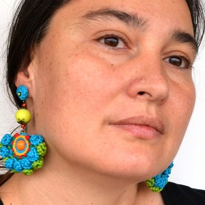 May include: A pair of colorful crocheted earrings with a flower design. The earrings are blue, green, yellow, orange, and red. The earrings are dangling from the woman's ears.