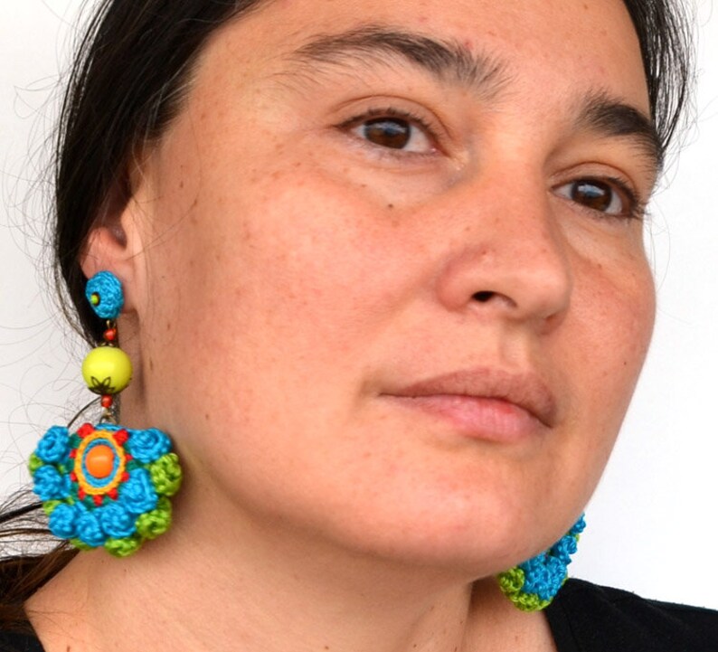 May include: A pair of colorful crocheted earrings with a flower design. The earrings are blue, green, yellow, orange, and red. The earrings are dangling from the woman's ears.