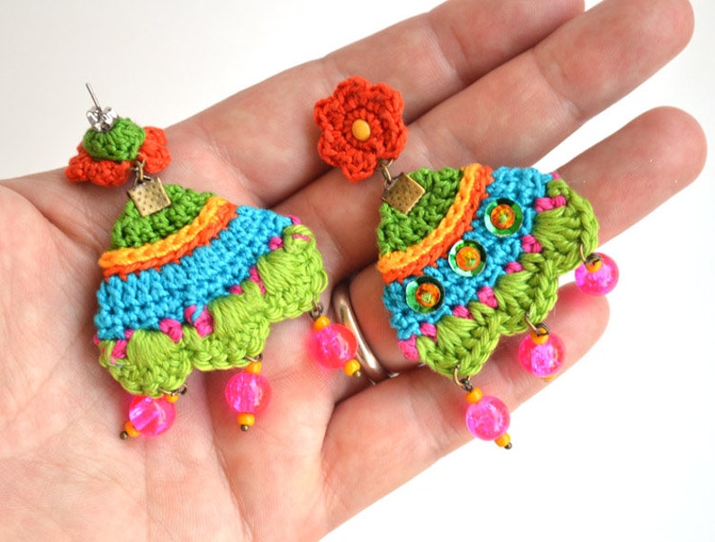 Puede incluir: Un par de pendientes de ganchillo coloridos con un dije de flor y cuentas rosadas. Los pendientes est&aacute;n hechos con hilo verde, azul, naranja y rosa.