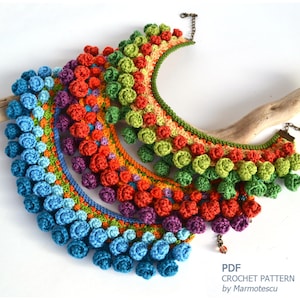May include: Three crocheted necklaces with colorful graduated pom-poms. The necklaces are made in shades of blue, purple, orange, and green. The necklaces are arranged in a cascading pattern, with the largest necklace at the bottom and the smallest necklace at the top.  PDF CROCHET PATTERN by Marmotescu.