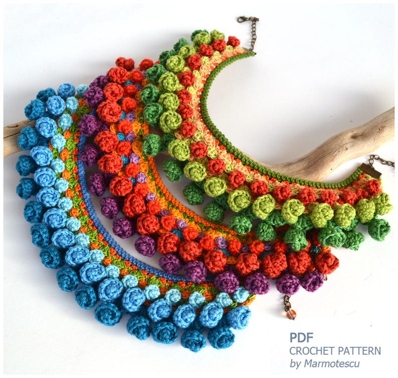 May include: Three crocheted necklaces with colorful graduated pom-poms. The necklaces are made in shades of blue, purple, orange, and green. The necklaces are arranged in a cascading pattern, with the largest necklace at the bottom and the smallest necklace at the top.  PDF CROCHET PATTERN by Marmotescu.