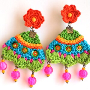Puede incluir: Un par de pendientes de ganchillo coloridos con un dije de flor, cuentas verdes, azules, rosas, naranjas y amarillas, y lentejuelas.