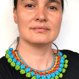 May include: A colorful crocheted necklace with a bright blue base and a row of green, orange, and purple crocheted flowers. The necklace is worn by a person with brown hair and brown eyes.