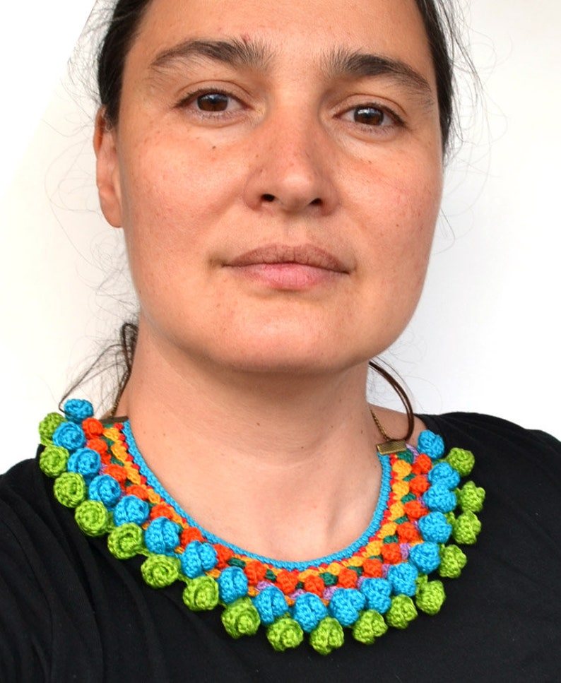 May include: A colorful crocheted necklace with a bright blue base and a row of green, orange, and purple crocheted flowers. The necklace is worn by a person with brown hair and brown eyes.
