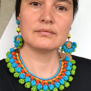 May include: A colorful crocheted necklace and matching earrings. The necklace is made of bright blue, green, orange, and yellow yarn and features a row of crocheted flowers. The earrings are also crocheted and feature a similar flower design.