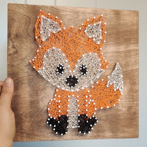 Könnte beinhalten: Ein Fadenkunst-Fuchs mit orangefarbenem, weißem und schwarzem Faden auf einem Holzgrund.