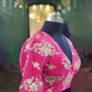 May include: A pink, short-sleeved blouse with gold embroidery. The blouse has a deep V-neckline and is fitted.