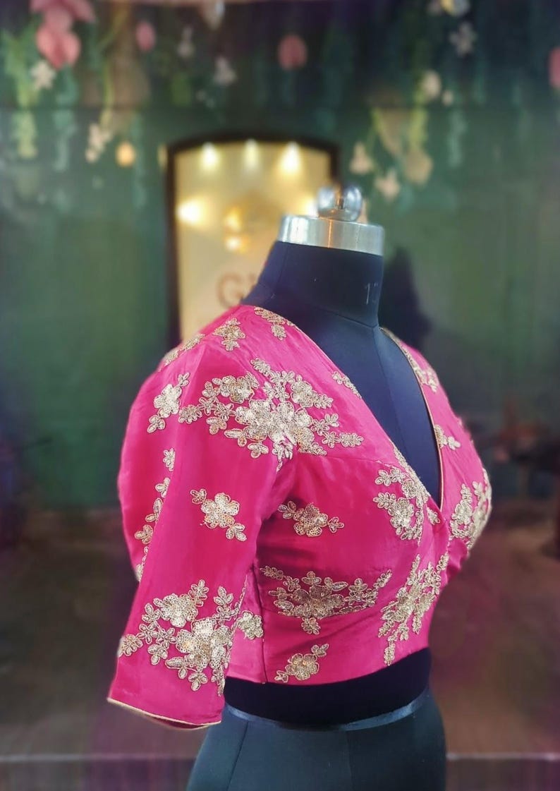 May include: A pink, short-sleeved blouse with gold embroidery. The blouse has a deep V-neckline and is fitted.