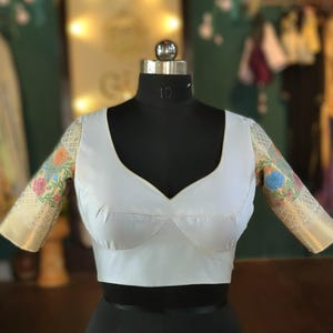 Puede incluir: Blusa blanca con escote en V profundo y mangas estampadas con flores. Las mangas presentan una base color crema con bordados florales coloridos. La blusa se exhibe en un maniquí negro.
