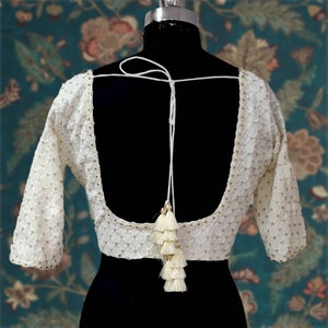 Puede incluir: Blusa blanca bordada con espalda baja y cierre de lazo con borla. La blusa tiene un patr&oacute;n delicado y est&aacute; hecha de una tela ligera.