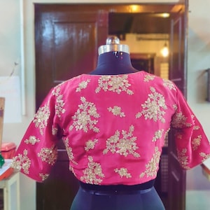 May include: A pink embroidered blouse with gold floral details. The blouse has short sleeves and a round neckline.