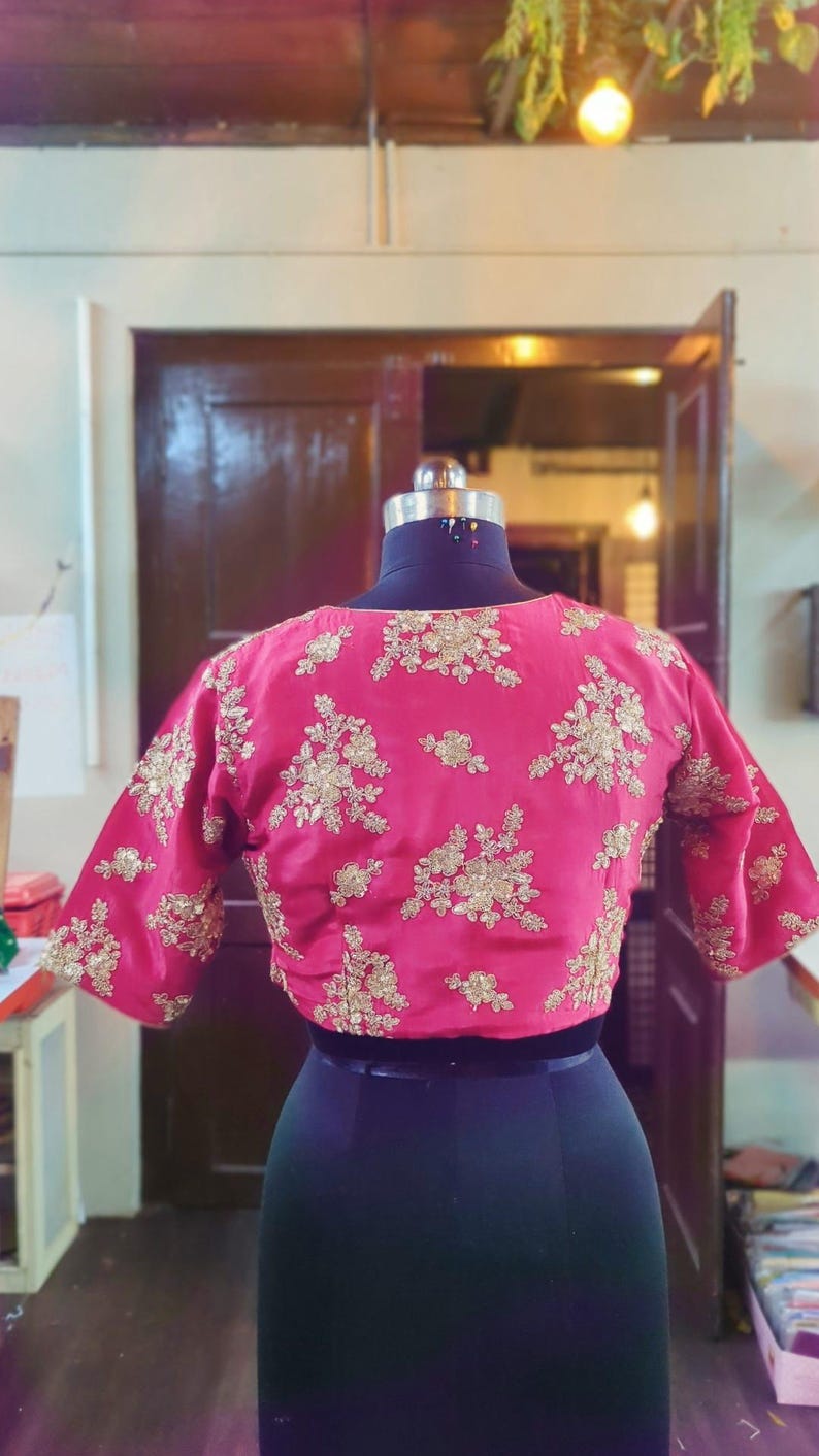 May include: A pink embroidered blouse with gold floral details. The blouse has short sleeves and a round neckline.