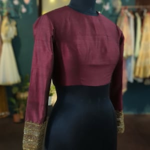 May include: A maroon blouse with long sleeves and gold embroidery on the cuffs. The blouse is on a black mannequin.