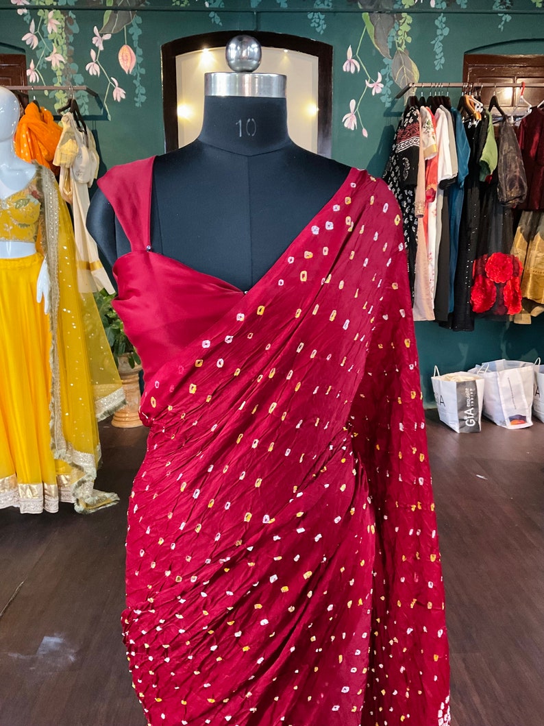 May include: A burgundy saree with a repeating pattern of small yellow and white dots, draped on a black mannequin. The saree's fabric is textured and appears to be silk or a similar material. The mannequin is in a boutique setting, with other garments visible in the background.