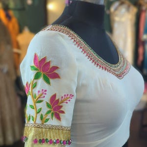 Puede incluir: Blusa blanca con flores de loto bordadas en rosa y verde en las mangas. El escote y los puños están adornados con intrincados adornos dorados, rojos y verdes. La blusa se exhibe en un maniquí negro.