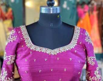 May include: A magenta-colored blouse with gold embroidery on the neckline and sleeves. The blouse is displayed on a black mannequin. The bottom of the blouse is paired with a matching skirt, also with gold embroidery. The outfit is likely part of a traditional Indian or South Asian ensemble.