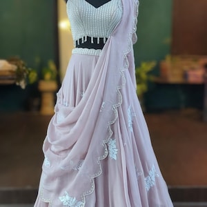 May include: A light pink, floor-length skirt with white floral embroidery and a scalloped hem. The skirt is paired with a silver sequined crop top with a beaded fringe.