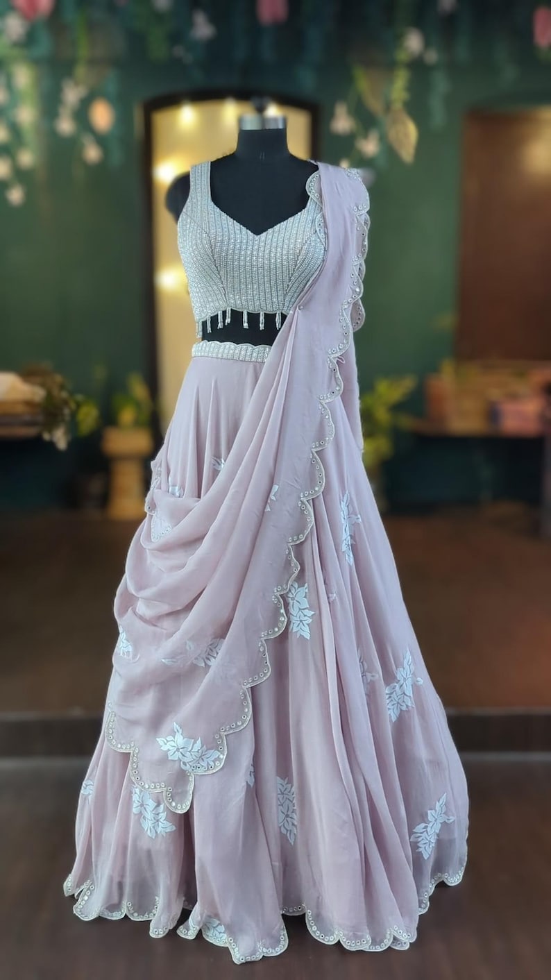 May include: A light pink, floor-length skirt with white floral embroidery and a scalloped hem. The skirt is paired with a silver sequined crop top with a beaded fringe.