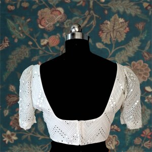May include: White embroidered blouse with a scoop neck and puff sleeves. The blouse is decorated with small, round mirrors.