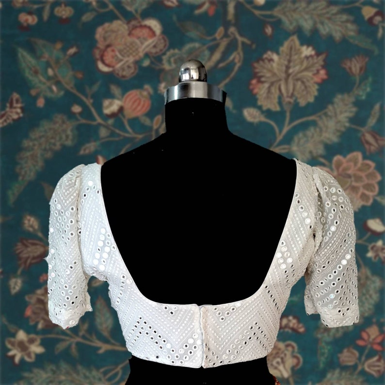 May include: White embroidered blouse with a scoop neck and puff sleeves. The blouse is decorated with small, round mirrors.